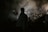 A person cloaked in dark clothing holds a large circular object resembling a religious monstrance. Smoke or mist swirls around, casting an ethereal and mysterious atmosphere.