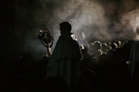 A person cloaked in dark clothing holds a large circular object resembling a religious monstrance. Smoke or mist swirls around, casting an ethereal and mysterious atmosphere.