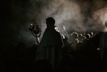 A person cloaked in dark clothing holds a large circular object resembling a religious monstrance. Smoke or mist swirls around, casting an ethereal and mysterious atmosphere.