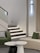 A modern and minimalistic interior featuring a staircase with illuminated steps, accompanied by a green armchair with a checkered pillow and a beige sofa with a green pillow. In the foreground, two round tables, one black and one white, stand out against the light tiled floor. A sleek, vertical light fixture mounted on the wall adds an artistic touch.