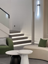 A modern and minimalistic interior featuring a staircase with illuminated steps, accompanied by a green armchair with a checkered pillow and a beige sofa with a green pillow. In the foreground, two round tables, one black and one white, stand out against the light tiled floor. A sleek, vertical light fixture mounted on the wall adds an artistic touch.