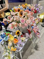 A display of intricately crocheted flowers in various colors including pink, yellow, blue, and purple arranged on a table. These crafted flowers resemble sunflowers, roses, and other floral varieties. Small decorative items, such as strawberries and animals, are also incorporated into the arrangement. A person stands nearby, partially visible.
