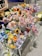 A display of intricately crocheted flowers in various colors including pink, yellow, blue, and purple arranged on a table. These crafted flowers resemble sunflowers, roses, and other floral varieties. Small decorative items, such as strawberries and animals, are also incorporated into the arrangement. A person stands nearby, partially visible.
