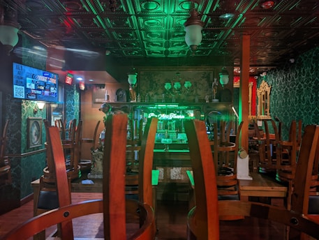 An empty bar with chairs stacked upside down on tables. The room is dimly lit with green and red neon lights reflecting off the ceiling and walls, which have intricate patterns. A neon sign behind the bar reads something partially visible. Decorative lamps hang from the ceiling, and there appears to be a TV screen on the left wall displaying various ads or events.