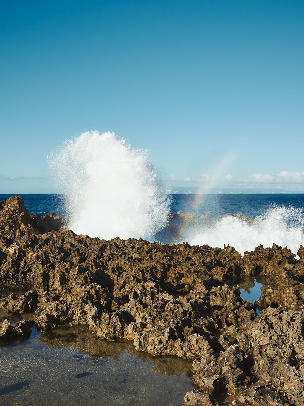 Una ola rompiendo en una orilla rocosa con un arco iris en el fondo