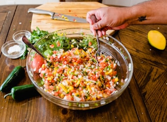 A large glass bowl is filled with fresh, colorful salsa made from diced tomatoes, onions, cilantro, and yellow corn. A hand with a tattoo is using a spoon and fork to mix the ingredients. On the wooden table, there are two green jalapeños, a half-sliced mango, a wooden chopping board with a knife, and small bowls of salt and pepper. Fresh cilantro is visible near the chopping board.