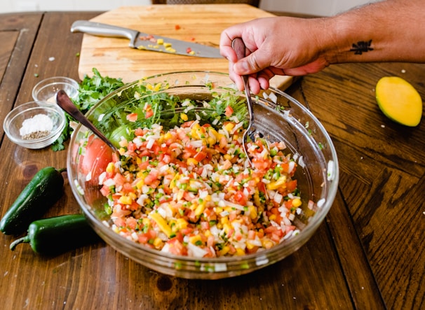A large glass bowl is filled with fresh, colorful salsa made from diced tomatoes, onions, cilantro, and yellow corn. A hand with a tattoo is using a spoon and fork to mix the ingredients. On the wooden table, there are two green jalapeños, a half-sliced mango, a wooden chopping board with a knife, and small bowls of salt and pepper. Fresh cilantro is visible near the chopping board.