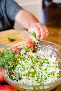 A person squeezing lime juice onto a bowl of chopped vegetables including onions and green peppers. The bowl is placed on a wooden surface with some whole tomatoes and another halved lime nearby.