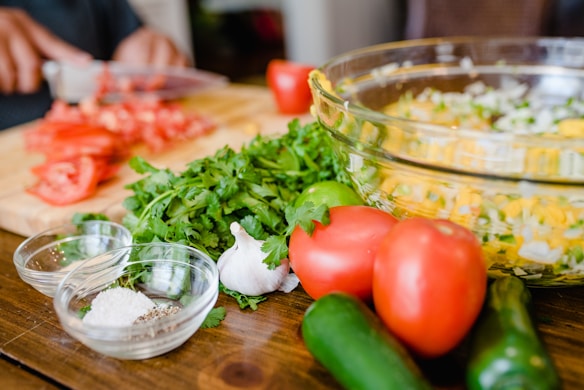 A wooden table is set with fresh ingredients including whole tomatoes, chopped tomatoes, garlic, cilantro, and cucumbers. There are also small glass bowls containing salt and pepper. A clear glass bowl filled with a mixture of chopped vegetables is visible, likely a salad or salsa. In the background, a person is chopping more tomatoes.