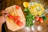 A person is slicing tomatoes on a wooden cutting board with various ingredients arranged around. Next to the board, there is a large glass bowl filled with a mixture of diced mango, onion, and cilantro. Fresh cilantro, limes, a whole mango, jalapeño peppers, and additional tomatoes are scattered on the table. Small bowls containing seasonings are also present.
