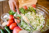 A hand is using a knife to add a portion of minced garlic to a large glass bowl filled with chopped onions and herbs. Surrounding the bowl on a wooden table are fresh ingredients including whole tomatoes, jalapeños, and pomegranates. Small bowls of ground spices and herbs are also positioned nearby.