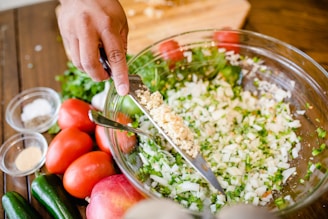 A hand is using a knife to add a portion of minced garlic to a large glass bowl filled with chopped onions and herbs. Surrounding the bowl on a wooden table are fresh ingredients including whole tomatoes, jalapeños, and pomegranates. Small bowls of ground spices and herbs are also positioned nearby.