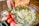 A hand is using a knife to add a portion of minced garlic to a large glass bowl filled with chopped onions and herbs. Surrounding the bowl on a wooden table are fresh ingredients including whole tomatoes, jalapeños, and pomegranates. Small bowls of ground spices and herbs are also positioned nearby.