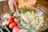 A hand is using a knife to add a portion of minced garlic to a large glass bowl filled with chopped onions and herbs. Surrounding the bowl on a wooden table are fresh ingredients including whole tomatoes, jalapeños, and pomegranates. Small bowls of ground spices and herbs are also positioned nearby.