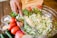 A hand is using a knife to add a portion of minced garlic to a large glass bowl filled with chopped onions and herbs. Surrounding the bowl on a wooden table are fresh ingredients including whole tomatoes, jalapeños, and pomegranates. Small bowls of ground spices and herbs are also positioned nearby.
