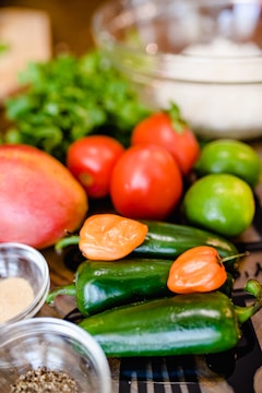 A collection of fresh ingredients including green jalapeños, orange habanero peppers, tomatoes, limes, and a mango. There is also a bunch of cilantro and two small bowls containing spices. A transparent glass bowl is partially visible in the background.