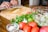 A person chops garlic on a wooden cutting board surrounded by fresh vegetables. A clear glass bowl contains finely chopped onions and herbs. The table includes tomatoes, cilantro, lime, and ingredients in a small glass bowl.