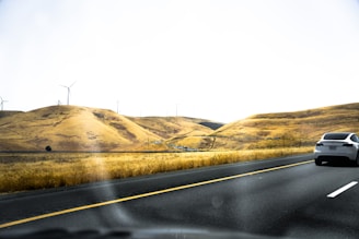 A sleek white car drives on a multi-lane highway through a hilly landscape with golden, dry grass. Wind turbines are scattered on the hills, indicating a wind farm. The sky is clear and bright, suggesting a sunny day.