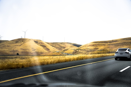 A sleek white car drives on a multi-lane highway through a hilly landscape with golden, dry grass. Wind turbines are scattered on the hills, indicating a wind farm. The sky is clear and bright, suggesting a sunny day.