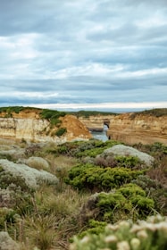 A landscape featuring rugged coastal terrain with green vegetation in the foreground. The scene includes limestone cliffs with stratified layers and scattered greenery on top. In the background, a narrow waterway is visible, framed by the cliffs, leading toward the ocean under an overcast sky filled with grey clouds.