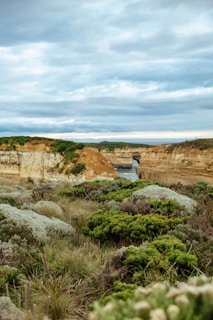 A landscape featuring rugged coastal terrain with green vegetation in the foreground. The scene includes limestone cliffs with stratified layers and scattered greenery on top. In the background, a narrow waterway is visible, framed by the cliffs, leading toward the ocean under an overcast sky filled with grey clouds.