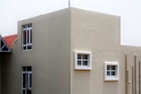 A modern building exterior with smooth beige walls and several windows, some framed in white. The building has a geometric design with a flat roof, and pipes run vertically along one side.