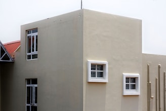 A modern building exterior with smooth beige walls and several windows, some framed in white. The building has a geometric design with a flat roof, and pipes run vertically along one side.