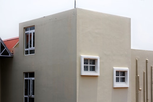 A modern building exterior with smooth beige walls and several windows, some framed in white. The building has a geometric design with a flat roof, and pipes run vertically along one side.