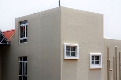 A modern building exterior with smooth beige walls and several windows, some framed in white. The building has a geometric design with a flat roof, and pipes run vertically along one side.