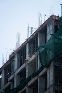 The image depicts a concrete building under construction with exposed rebars extending from the top. Green safety netting is draped along the sides of the structure. The concrete pillars and floors are visible, and the general skeletal framework of the building is prominent.