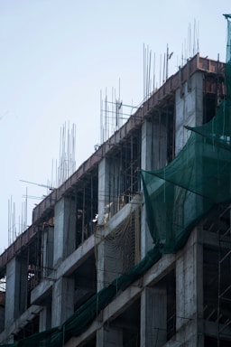 The image depicts a concrete building under construction with exposed rebars extending from the top. Green safety netting is draped along the sides of the structure. The concrete pillars and floors are visible, and the general skeletal framework of the building is prominent.