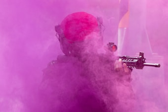 A person in tactical gear holds a rifle, obscured by thick pink smoke. The scene is dynamic, with contrasting colors and a sense of action.