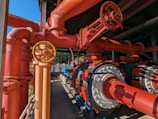 An intricate network of large red and orange industrial pipes and valves, along with metallic components. The system appears robust and well-constructed, with shadows and sunlight casting dynamic patterns on the surfaces. A pathway runs alongside, suggesting accessibility for maintenance.
