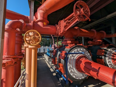 An intricate network of large red and orange industrial pipes and valves, along with metallic components. The system appears robust and well-constructed, with shadows and sunlight casting dynamic patterns on the surfaces. A pathway runs alongside, suggesting accessibility for maintenance.