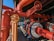 An intricate network of large red and orange industrial pipes and valves, along with metallic components. The system appears robust and well-constructed, with shadows and sunlight casting dynamic patterns on the surfaces. A pathway runs alongside, suggesting accessibility for maintenance.