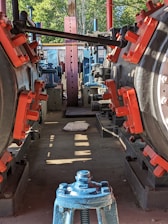 A mechanical setting with large industrial machinery featuring metal components, painted in red and blue. The machinery is aligned in a row, creating a corridor effect. Sunlight casts shadows on the concrete floor. A pillow is placed on the floor in the middle of the pathway. In the background, greenery is visible outside the structure.