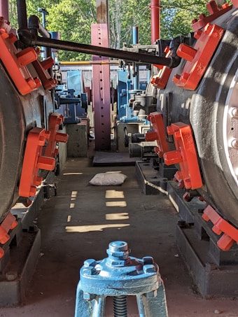 A mechanical setting with large industrial machinery featuring metal components, painted in red and blue. The machinery is aligned in a row, creating a corridor effect. Sunlight casts shadows on the concrete floor. A pillow is placed on the floor in the middle of the pathway. In the background, greenery is visible outside the structure.