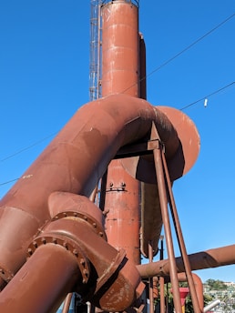A series of large, rust-colored industrial pipes with flanges and bolts dominate the scene. The pipes are positioned against a clear blue sky, giving a sense of size and industrial functionality.