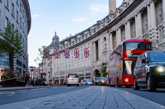 A bustling street scene features iconic British elements, including a notable red double-decker bus traveling alongside other vehicles. Union Jack flags are prominently displayed, adding a nationalistic touch. The architecture is grand with classical columns and intricate details. People are walking along the sidewalks, contributing to the lively atmosphere.