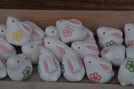 A collection of ceramic or porcelain rabbit figurines, predominantly white with pink inner ears and small red dots for eyes. Each rabbit is adorned with floral patterns in various colors such as yellow, green, and blue. They are positioned on a light wooden surface, adding a natural and cozy atmosphere.
