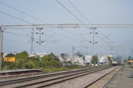 A railway track with overhead electric lines stretches into the distance, surrounded by greenery and urban buildings in the background. The platform sign reads 'Dhandera' in multiple scripts. The sky is clear, and a train is visible approaching along the tracks.