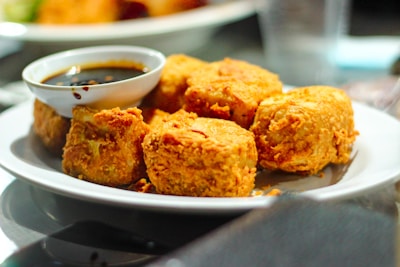 A plate of golden-brown fried tofu served with a small bowl of dark soy sauce on the side. The tofu pieces are crispy and appear appetizing, arranged neatly on a white plate.