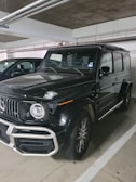 A black, luxury SUV with a bold front grille and large, round headlights is parked in a dimly lit indoor parking garage. The vehicle has a boxy and robust design, characteristic of high-end off-road vehicles. Dirt can be seen on the tires, suggesting recent use.