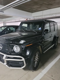 A black, luxury SUV with a bold front grille and large, round headlights is parked in a dimly lit indoor parking garage. The vehicle has a boxy and robust design, characteristic of high-end off-road vehicles. Dirt can be seen on the tires, suggesting recent use.
