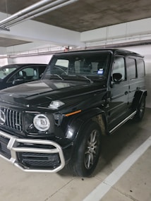 A black, luxury SUV with a bold front grille and large, round headlights is parked in a dimly lit indoor parking garage. The vehicle has a boxy and robust design, characteristic of high-end off-road vehicles. Dirt can be seen on the tires, suggesting recent use.