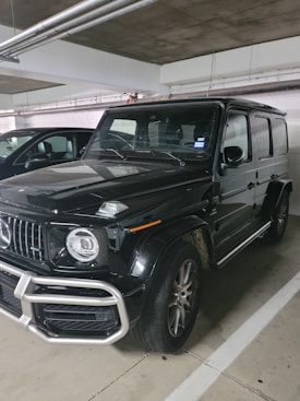 A black, luxury SUV with a bold front grille and large, round headlights is parked in a dimly lit indoor parking garage. The vehicle has a boxy and robust design, characteristic of high-end off-road vehicles. Dirt can be seen on the tires, suggesting recent use.