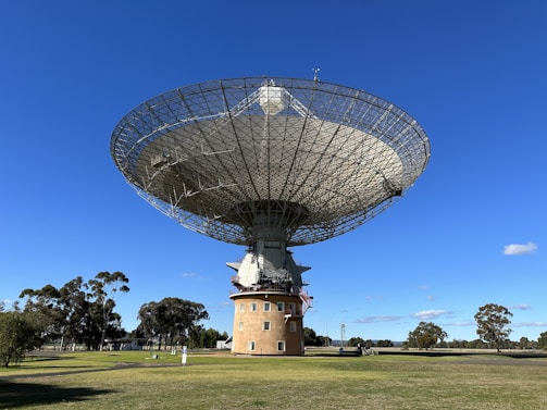 A large radio telescope with a parabolic dish structure stands prominently in a grassy field. The dish is mounted on a cylindrical base with a few small windows. Surrounding the structure, several trees are spread across the landscape under a clear blue sky.