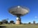 A large radio telescope with a parabolic dish structure stands prominently in a grassy field. The dish is mounted on a cylindrical base with a few small windows. Surrounding the structure, several trees are spread across the landscape under a clear blue sky.