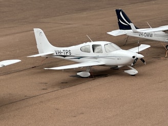 A small white aircraft is parked on an airfield with markings 'VH-TPS' and 'Cirrus SR20' visible on its side. The aircraft is secured with ropes attached to its wings. Another aircraft is partially visible in the background, marked 'VH-OWW.' The airfield surface is textured and light brown in color.
