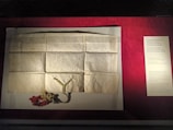 A historical document is displayed on a flat surface, consisting of aged, creased paper with handwritten script. Below the document, a decorative seal with green and red yarn is attached. To the right, a printed description on a board provides detailed context about the document, which appears to relate to a privilege issued by a king in 1433.